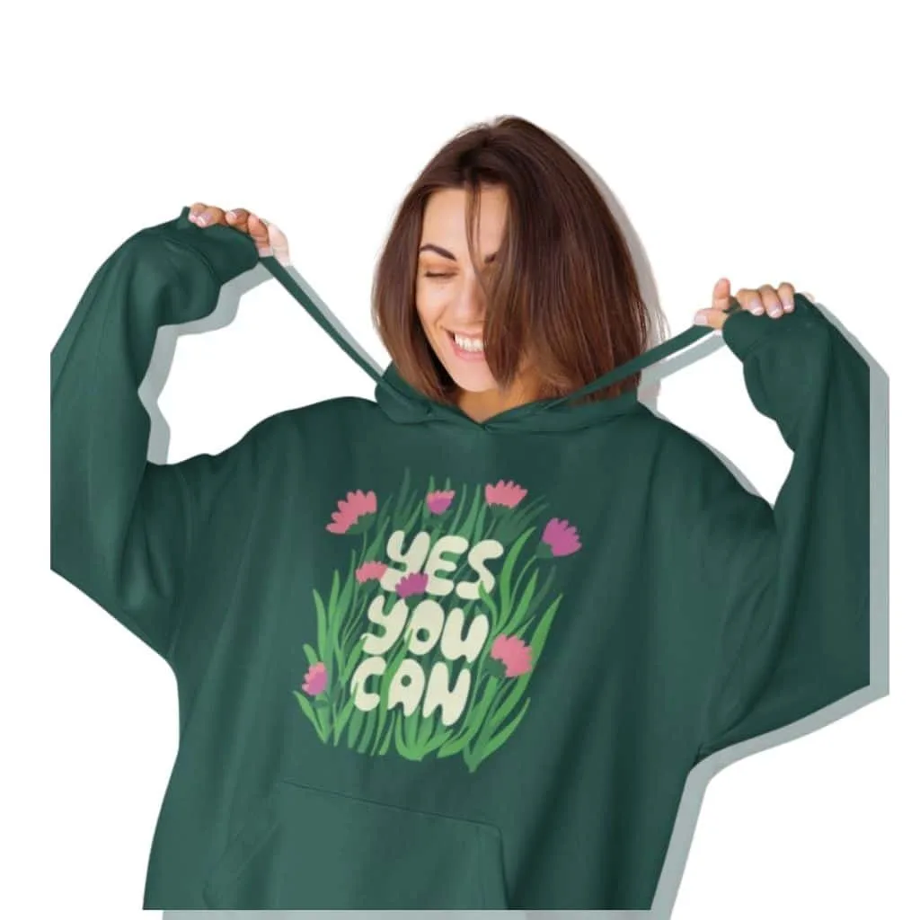A woman smiling while wearing a green hoodie featuring the slogan "YES YOU CAN" with floral graphics.