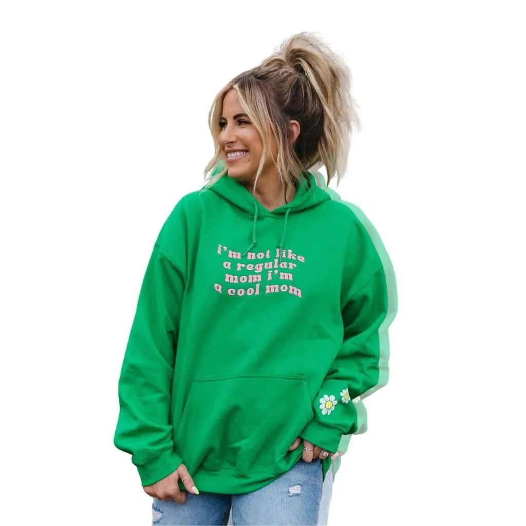 A woman wearing a bright green hoodie that reads "i’m not like a regular mom i’m a cool mom," smiling and posing.