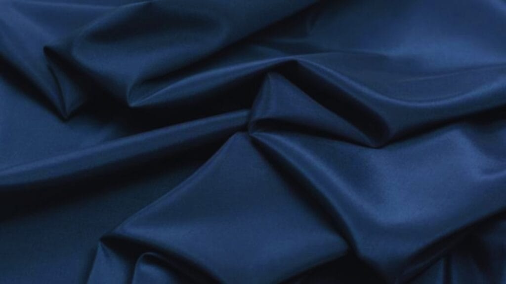 A close-up of dark blue fabric with smooth, shiny texture draped into folds. The material creates highlights and shadows, giving a three-dimensional, flowing appearance.