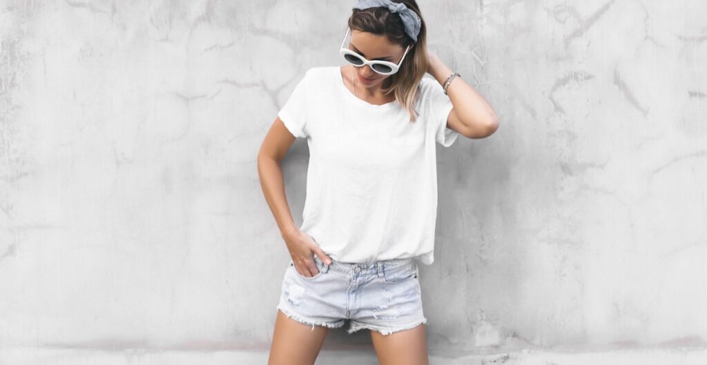 A young woman wearing a activewear white t-shirt and distressed denim shorts poses against a textured gray wall, accessorized with white sunglasses and a hair scarf.
