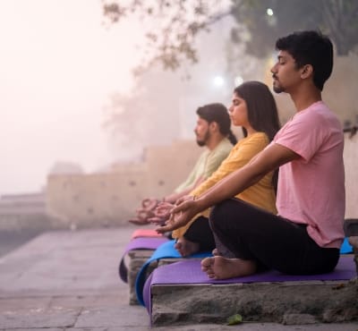 Three individuals meditating on yoga mats by a riverside in the early morning mist.