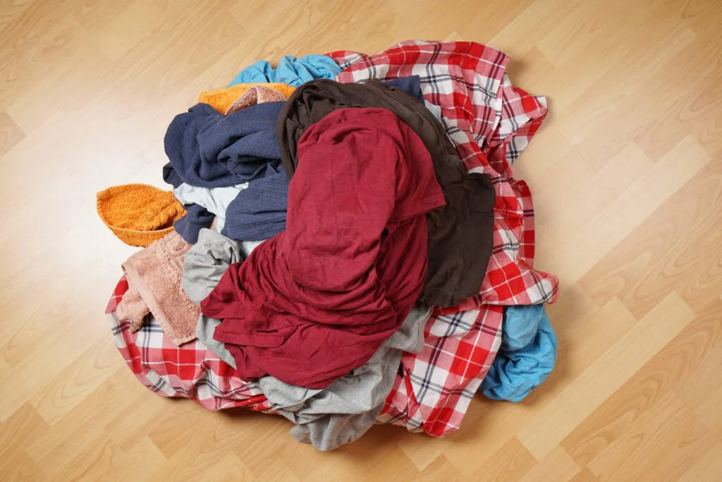 A pile of wrinkled clothes of various colors and textures, including red, gray, plaid, and blue, resting on a wooden floor.