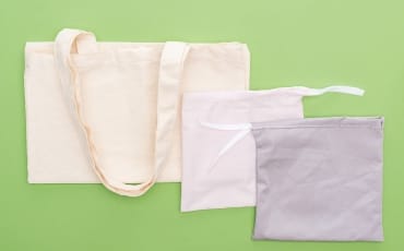A collection of cotton bags, including a large beige tote bag and two smaller pouches in light pink and gray, situated against a green background.