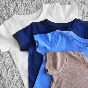 A stack of cotton t-shirts in various colors: white, dark blue, light blue, and brown, resting on a gray textured surface.