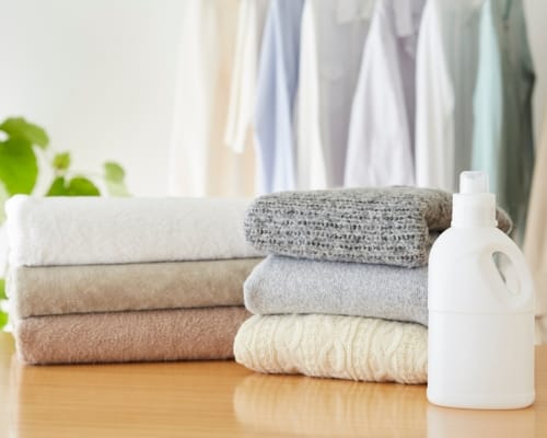 A neatly stacked pile of folded cotton towels next to a white laundry detergent bottle, with hanging clothes in the background.