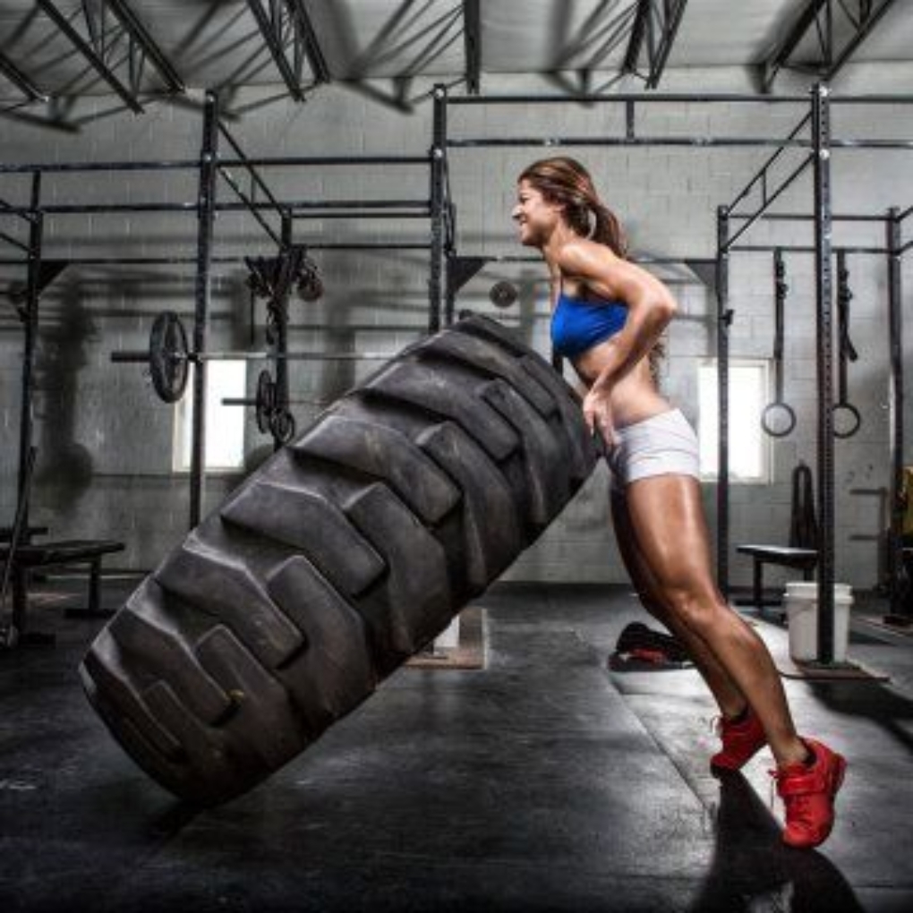 A muscular woman in a blue sports bra and white shorts is lifting a large tire in a gym setting, with exercise equipment visible in the background. Gym wear Guid