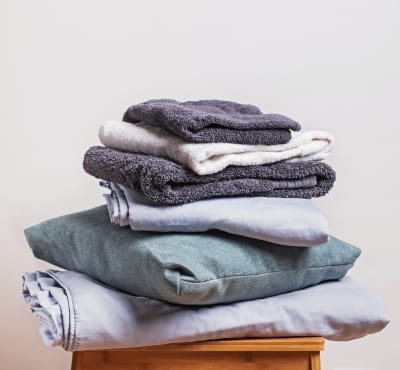 A neatly stacked pile of soft cotton towels and blankets in various shades, including gray, white, and light blue, resting on a wooden surface. products from the cotton