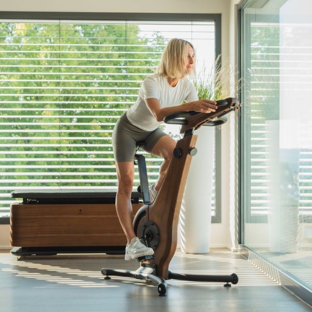 A woman with blonde hair is exercising on a modern wooden stationary bike in a bright room with large windows and blinds. She is dressed in a white shirt and gray shorts, focusing intently on her workout.
Gym wear guid
