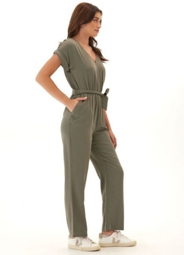 A woman stands confidently in a relaxed olive green jumpsuit with short sleeves, a V-neckline, and a cinched waist. She wears white sneakers and poses with her hands in the jumpsuit's pockets.