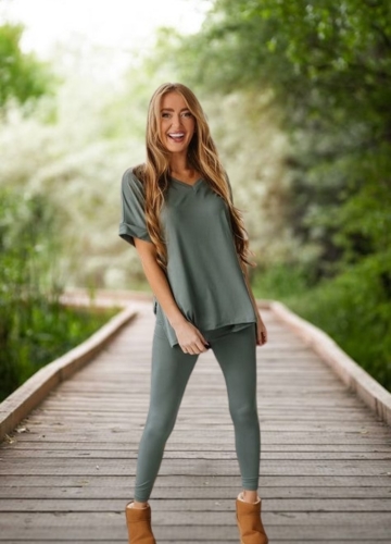 A woman stands on a wooden walkway surrounded by greenery, wearing an olive green loungewear set with a loose-fitting top and leggings, paired with tan ankle boots. She is smiling and looks relaxed.