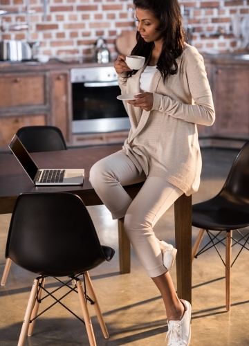 A woman in loungewear sits at a table, sipping tea or coffee while working on a laptop in a cozy kitchen.