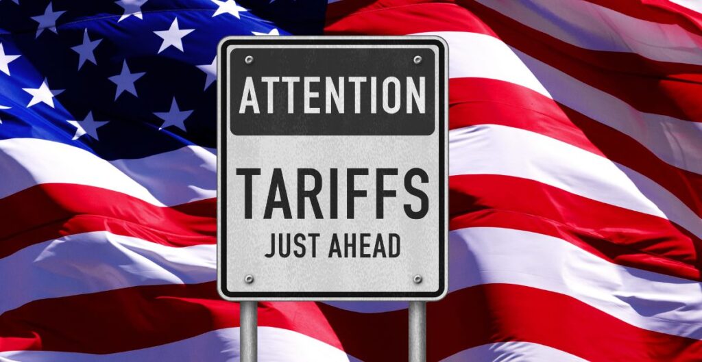 A road sign reads "ATTENTION TARIFS JUST AHEAD" in front of a waving American flag background.