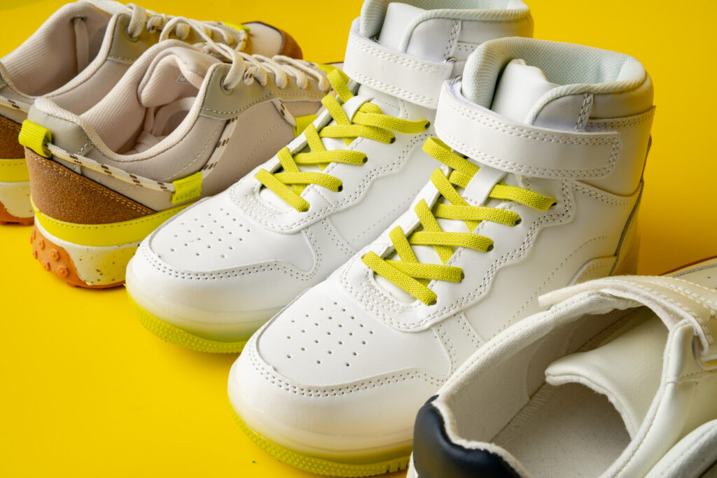 A collection of stylish sneakers displayed on a bright yellow background. The foreground features white high-top sneakers with yellow laces and a modern design, while the background has low-top sneakers in neutral tones with a mix of textures.