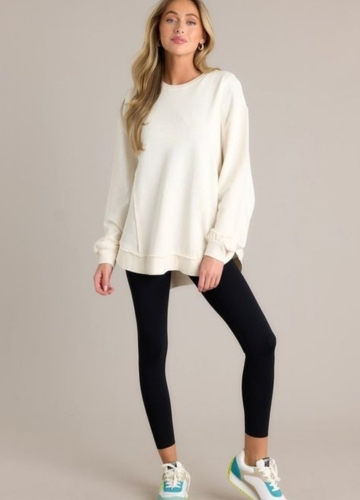 A model wearing an oversized cream sweatshirt, black leggings, and colorful sneakers, posing against a neutral background.