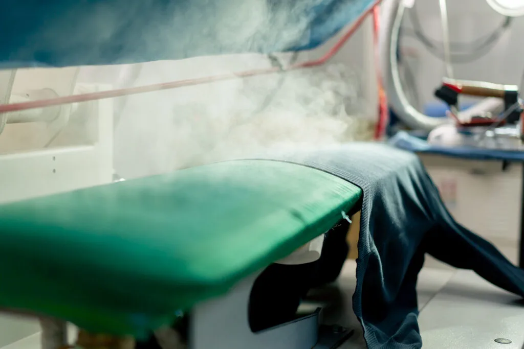 A close-up view of a garment fabric being prepared with steam, featuring a green ironing board and a piece of clothing draped over it, with steam rising in the background.