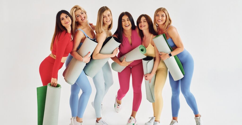 A group of six women in colorful athletic wear, each holding yoga mats, posing together and smiling joyfully.