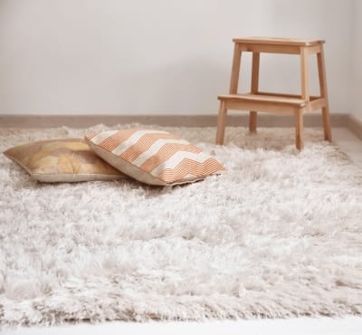 A cozy room featuring a plush white rug with two decorative pillows—one with a striped pattern and the other with a geometric design. A small wooden step stool is in the background.