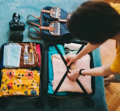Person packing a suitcase with clothing and travel essentials such as a camera, toiletries, and face masks.