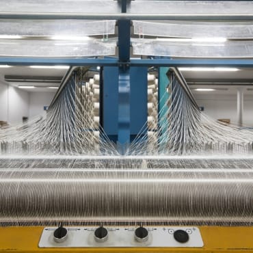 Close-up of a cotton weaving machine in a textile factory, showing multiple spools of yarn aligned horizontally and threads being woven into fabric, with spindles and guiding mechanisms visible.