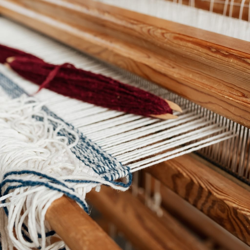 A close-up of a weaving loom with partially woven fabric, a bundle of red yarn, and a wooden comb or tool resting on the loom.