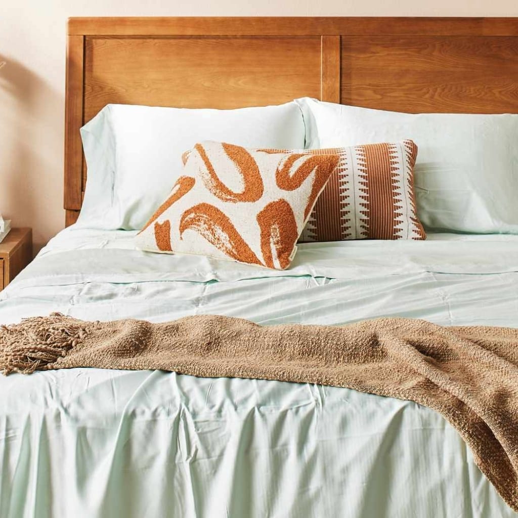 A cozy bed with a light green bedspread, decorative pillows including one with a rust-colored abstract pattern and another with a striped geometric design, a beige throw blanket, and a wooden headboard.