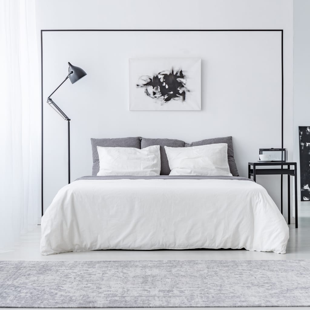 A minimalist bedroom with a white bed featuring gray and white pillows, a black framed artwork above, and a side table with a small decoration. A black adjustable floor lamp stands next to the bed, and the room has a clean, modern aesthetic with mostly white and black tones.