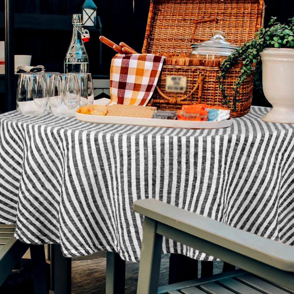 A close-up view of a table covered with a black and white striped tablecloth, set with four empty glasses, a small plate with a cookie and some bread, and some packaged snacks. In the background, there is a woven picnic basket with a lid and a checkered cloth sticking out, a glass water bottle, and a potted plant with green foliage.