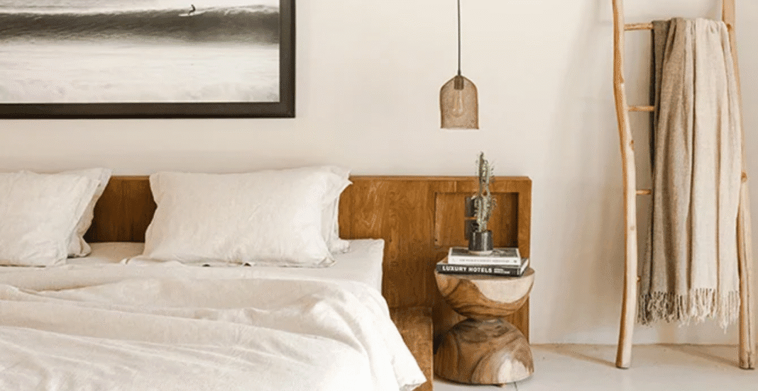 A cozy bedroom featuring a white bed with plush pillows, a wooden nightstand with a stack of books and a small potted plant, and a rustic wooden ladder with a blanket hanging on it, all in neutral, earthy tones.