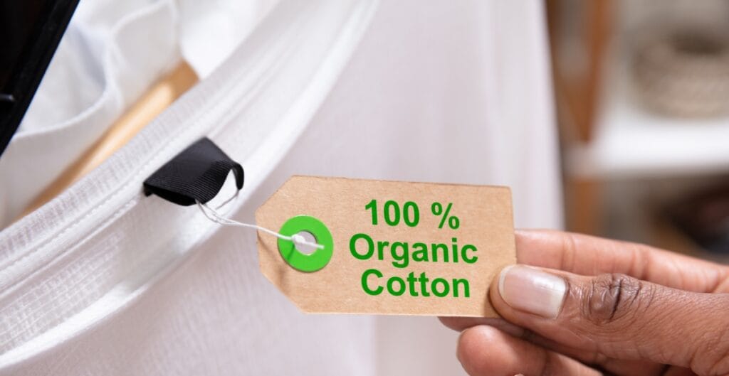 A hand holding a brown tag that reads "100% Organic Cotton" attached to a white fabric product with a black anchoring loop