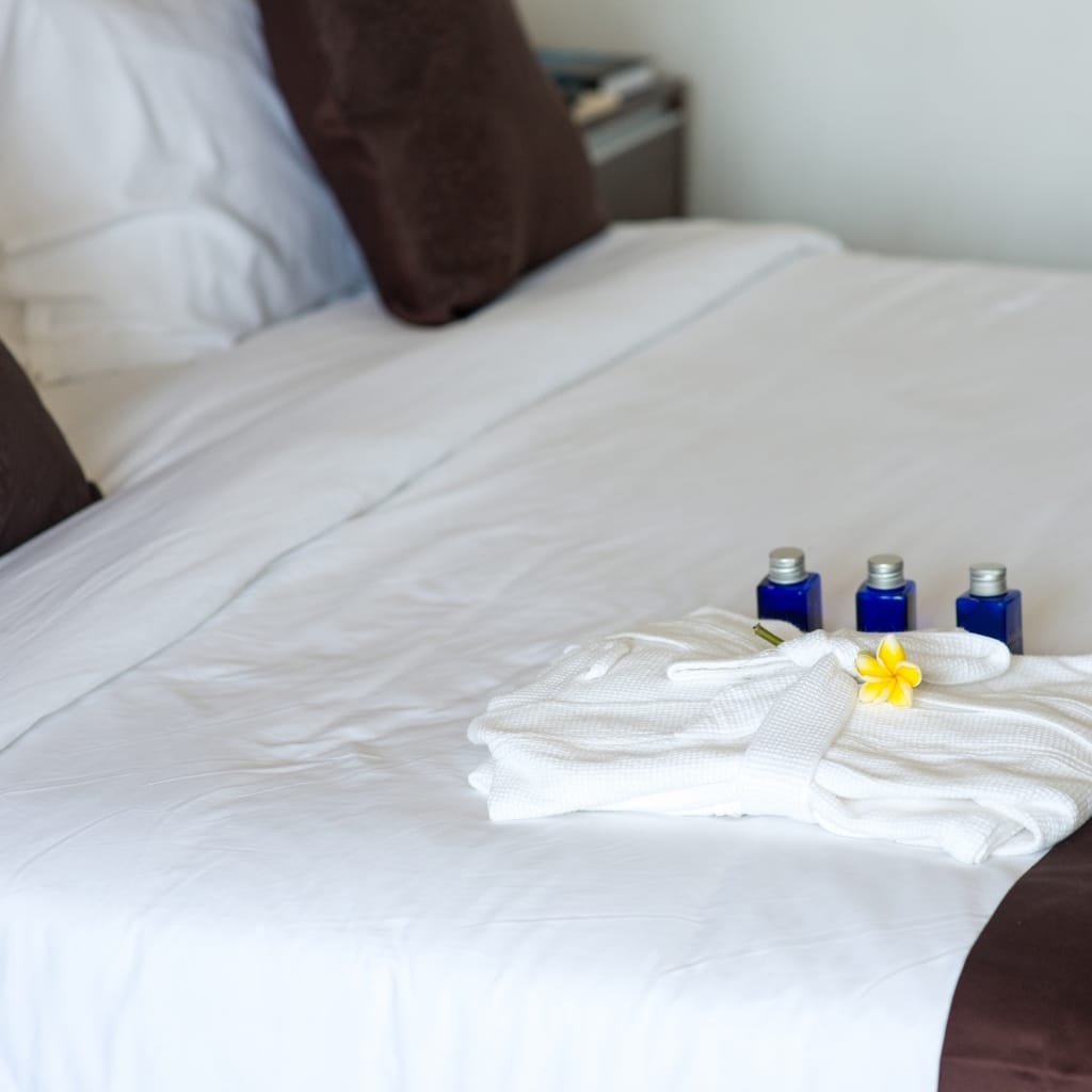 A neatly made bed with white bedding, dark brown pillows, and a stack of white towels adorned with a yellow flower, alongside three small blue bottles placed on the bed.