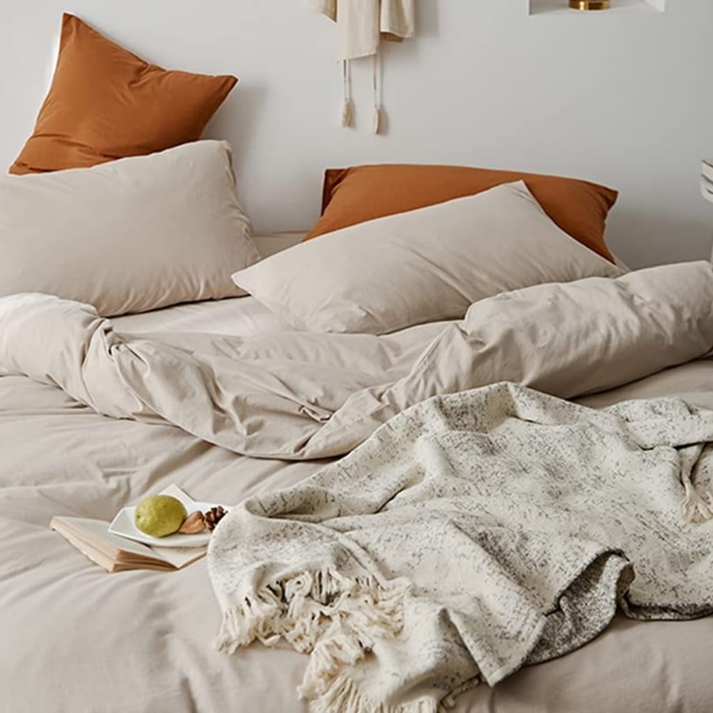 A cozy, inviting bed with white and beige bedding, a textured throw blanket, an open book, and a plate with a pear, nuts, and a pine cone on top of the bed.