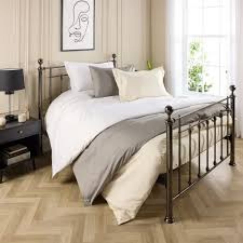A stylish bedroom featuring a metal-framed bed with white bedding, multiple pillows, and a gray throw blanket. There is a black bedside table with a lamp and a window with sheer curtains letting in natural light.