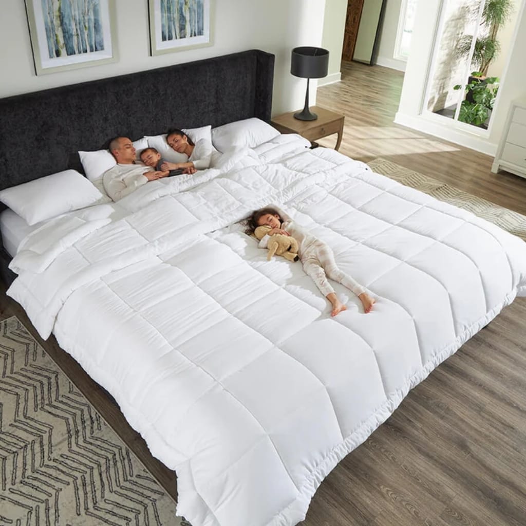 A family of four, including two adults and two children, lying on a large, white, neatly made bed in a bright, modern bedroom. The parents and one child are close together at one end of the bed, while a girl sleeps alone at the other end holding a teddy bear.