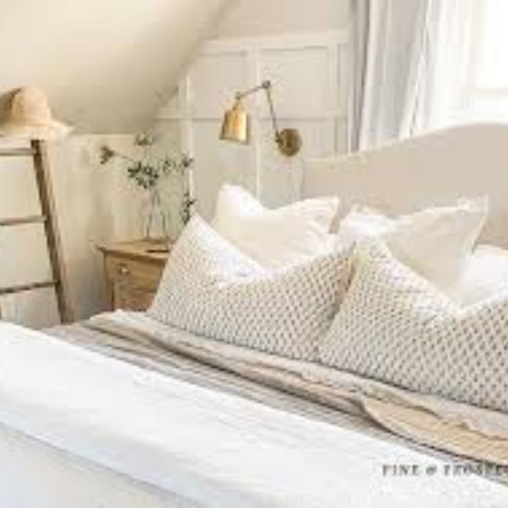 A cozy, well-lit bedroom corner featuring a bed with patterned pillows, a wooden bedside table with a vase of greenery, and a wall-mounted lamp. Light curtains and a window add to the airy ambiance.