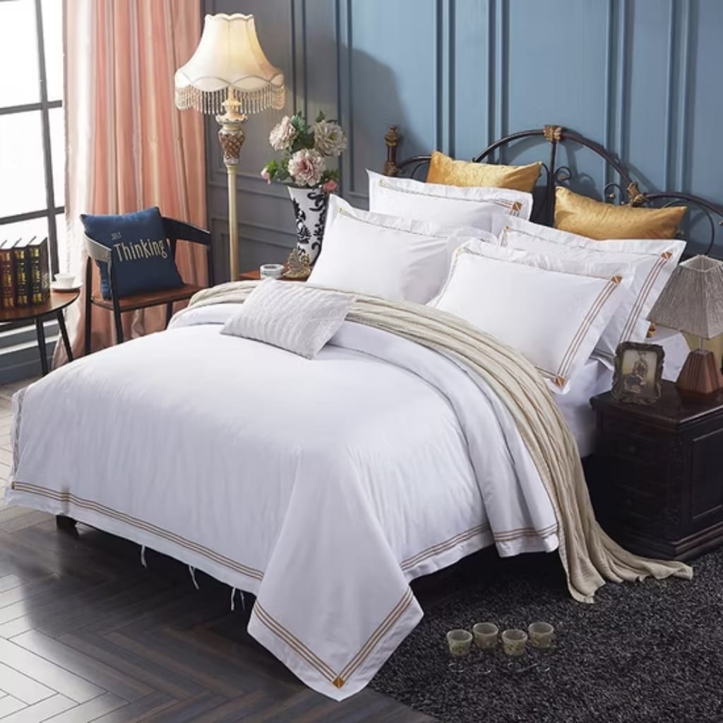 A luxurious bedroom featuring a large bed with white bedding, multiple pillows, a gold throw, and a dark wooden nightstand with a lamp, framed photo, and decorative items. There is a dark carpet, a chair with a book, a flower vase, and a vintage lamp against a teal paneled wall.
