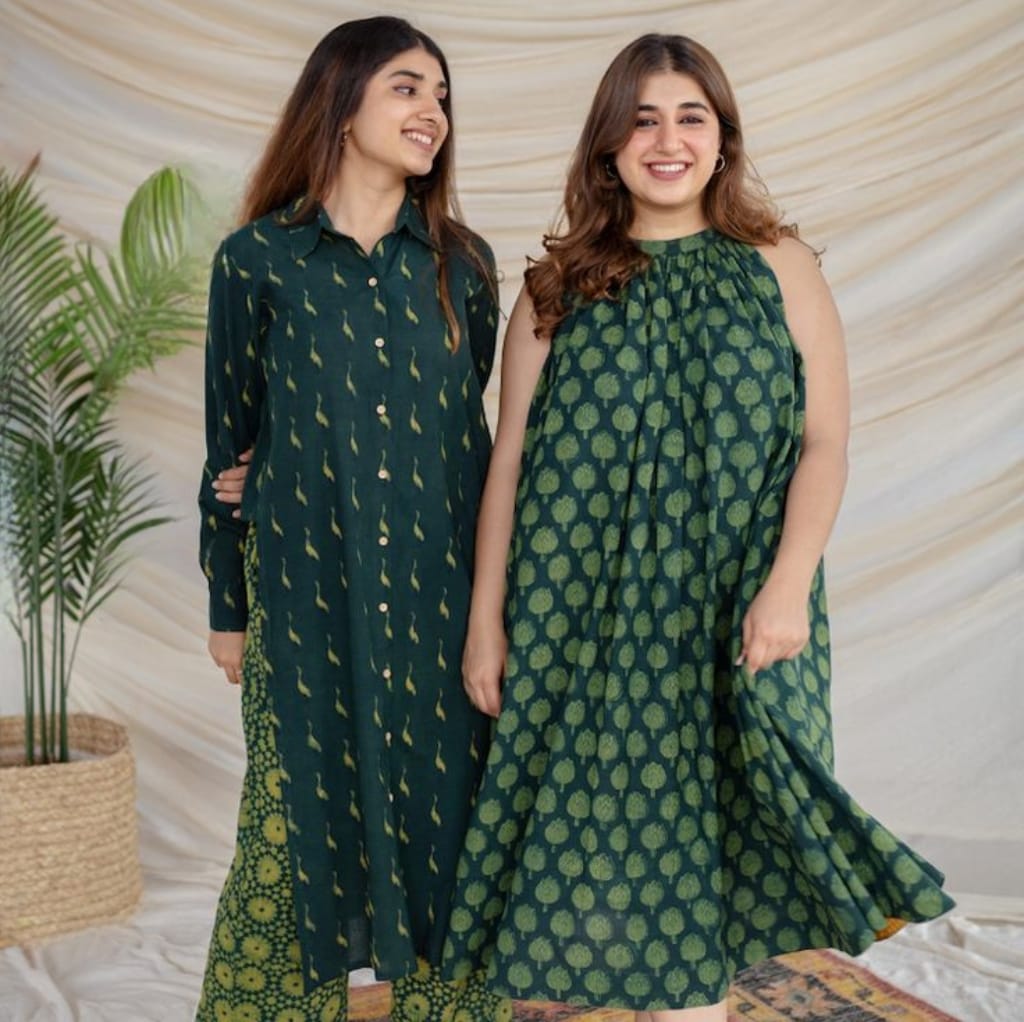 Two women standing together, wearing stylish cotton batiste fabric dresses, smiling, with a neutral background and decorative plants.