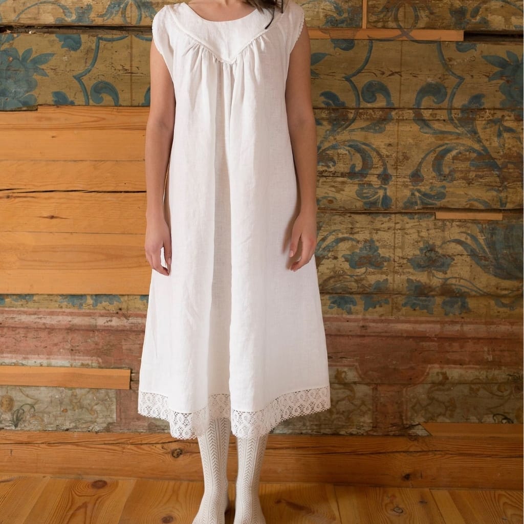 A woman wearing a long, sleeveless white dress with lace detail at the hem, standing against a rustic wooden wall with painted decorative patterns.