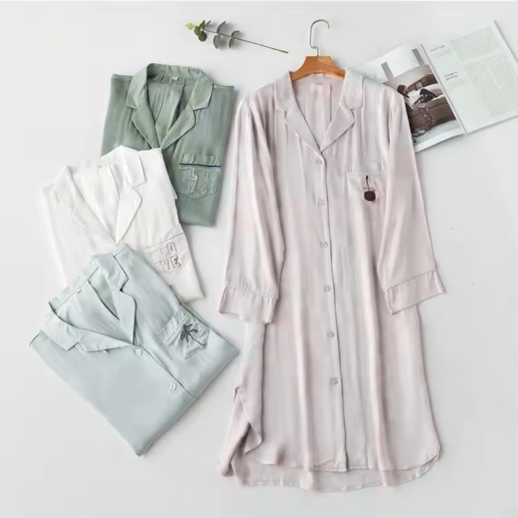 A flat lay of four silk pajamas in pastel and neutral colors, along with an open magazine, a small branch with leaves, and a hanger, all arranged on a white surface.