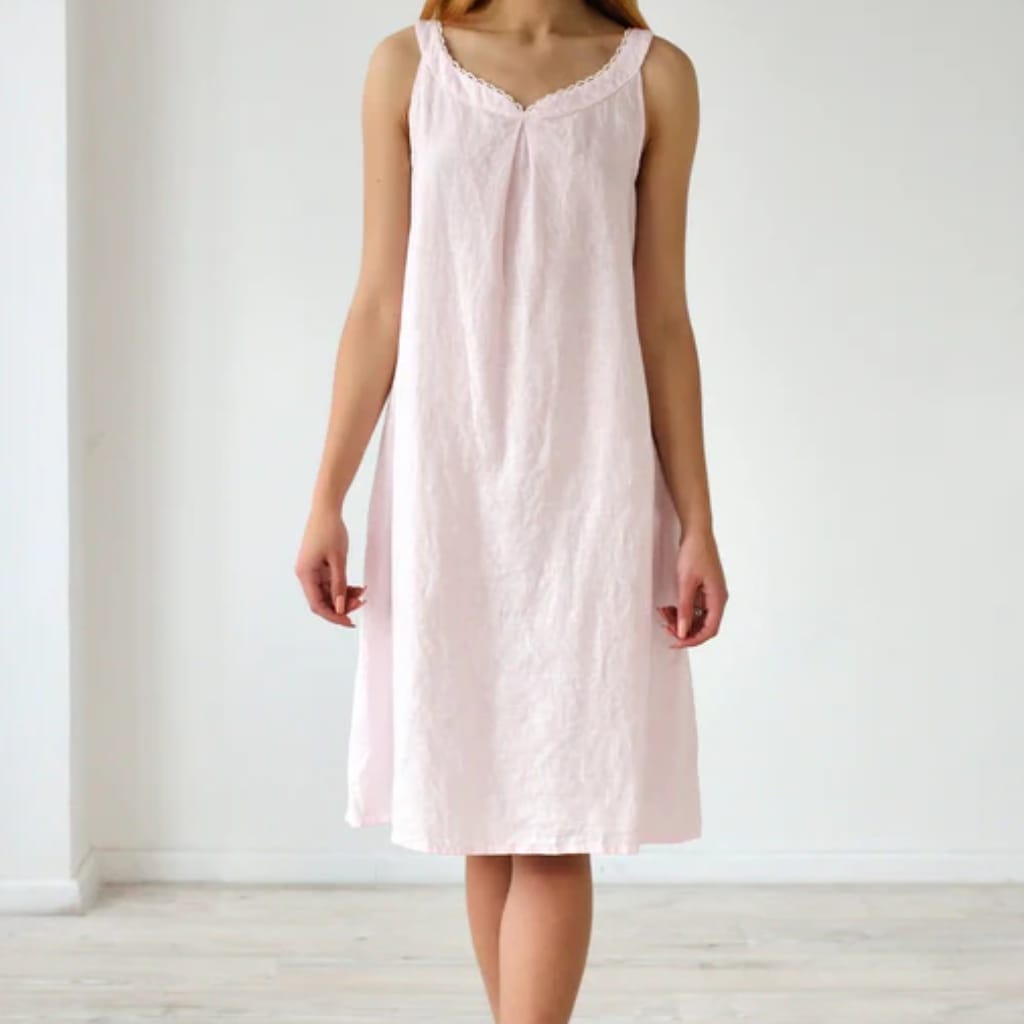 A woman wearing a light pink, sleeveless, knee-length nightgown with a V-neckline and lace trim, standing against a plain white wall.