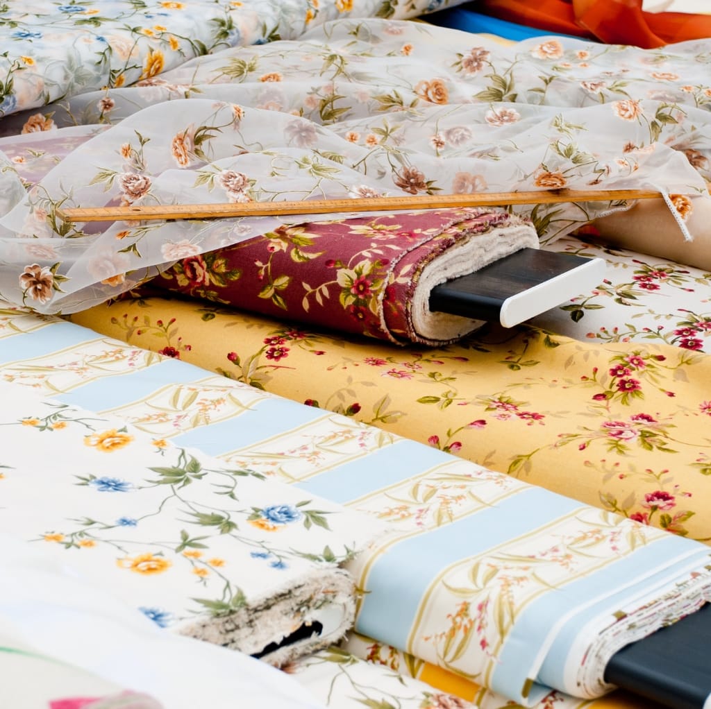 A close-up photograph of various patterned fabrics, including floral and striped designs, showcasing different types of textiles stacked and laid out, with a measuring stick resting across the fabrics.
