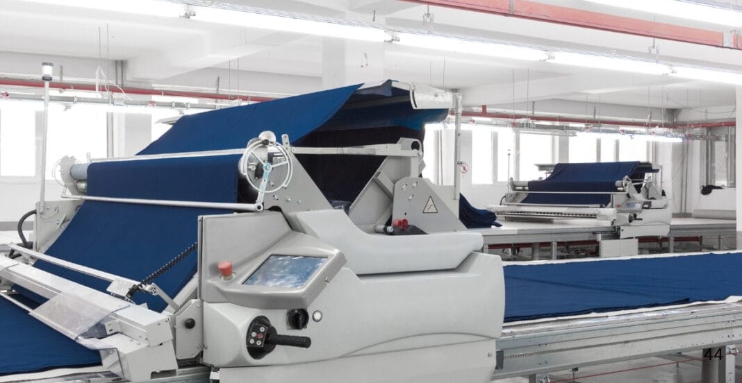 Automated textile manufacturing machine weaving blue fabric in a modern factory setting. This image depicts a high-tech textile production line, where automated machinery is weaving or processing blue fabric. The environment appears clean and well-lit, indicative of a contemporary textile manufacturing facility, showcasing the industrial scale and automation involved in American-made textiles.