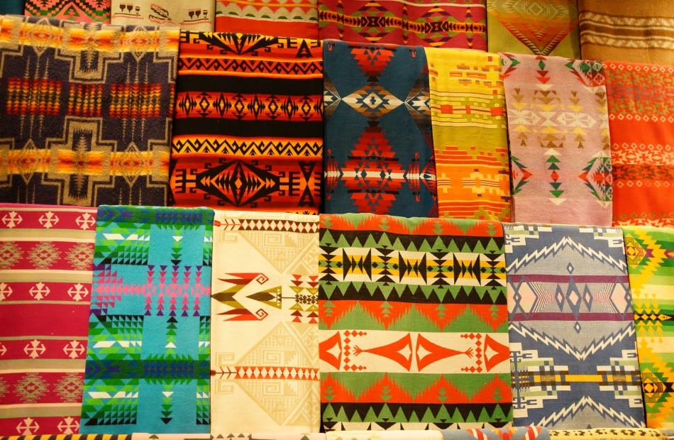 A colorful collection of various woven textiles with geometric patterns, including stripes, diamonds, and intricate motifs in vibrant shades of red, yellow, blue, green, orange, and beige.