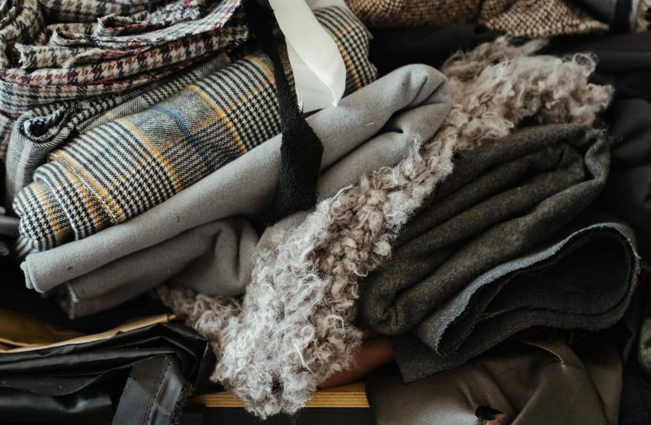 A pile of folded winter clothing and textiles, including plaid wool scarves, gray and dark-colored sweaters, and fluffy fleece-lined garments.