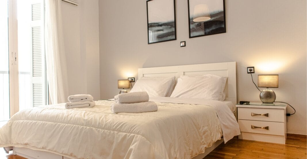 A neatly made bed with white bedding, white towels placed on top, flanked by matching bedside tables with lamps, and a balcony with white curtains on the left.