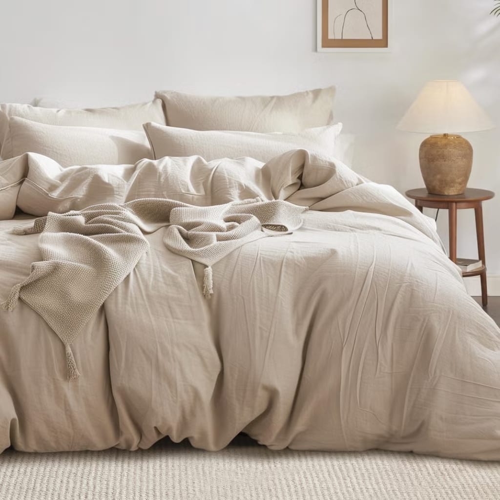 A cozy, neatly made bed with off-white, textured bedding and multiple pillows. A soft blanket is casually draped over the bed. To the right, there is a wooden side table with a large, earthy-toned lamp, and a simple, minimalistic framed line drawing on the wall behind.