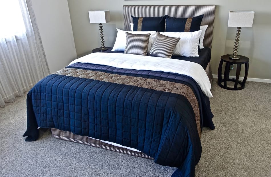 A neatly made bedroom featuring a bed with a dark blue quilted comforter, several decorative pillows, and two black nightstands with matching modern lamps, situated in a bright room with light-colored walls and carpet.
