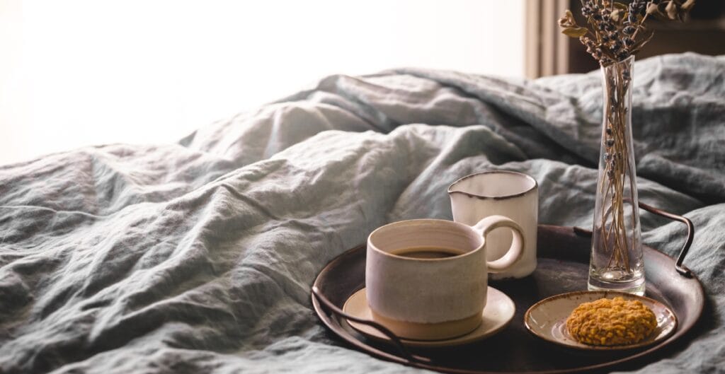 A cozy breakfast tray with a cup of coffee, a small mug, a cookie on a plate, and a vase with dried flowers, placed on a rumpled bed with soft, natural lighting.