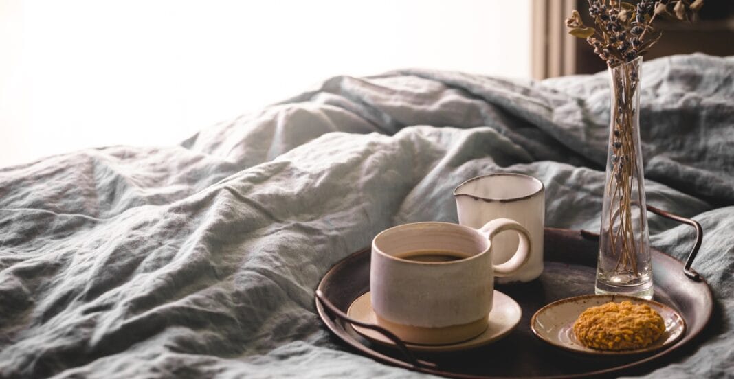 A cozy breakfast tray with a cup of coffee, a small mug, a cookie on a plate, and a vase with dried flowers, placed on a rumpled bed with soft, natural lighting.