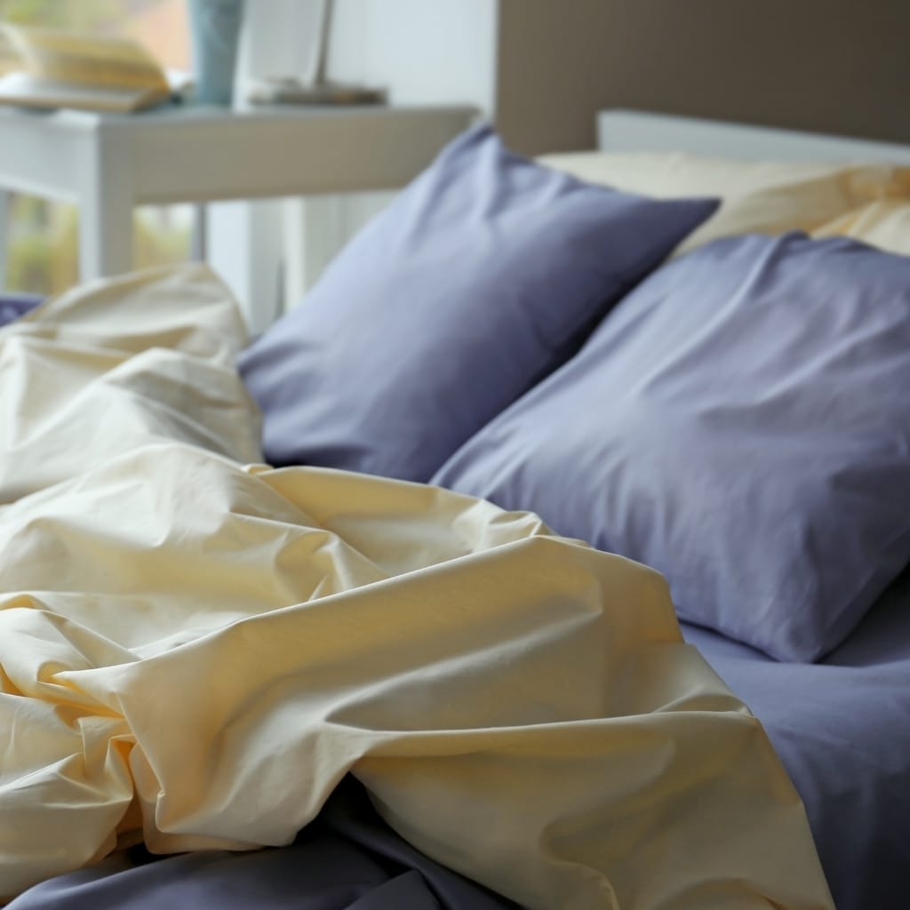 A cozy bedroom scene with a neatly made bed, featuring purple pillows, soft yellow and purple bed sheets, and a white bedside table with books, in front of a window with natural light streaming in