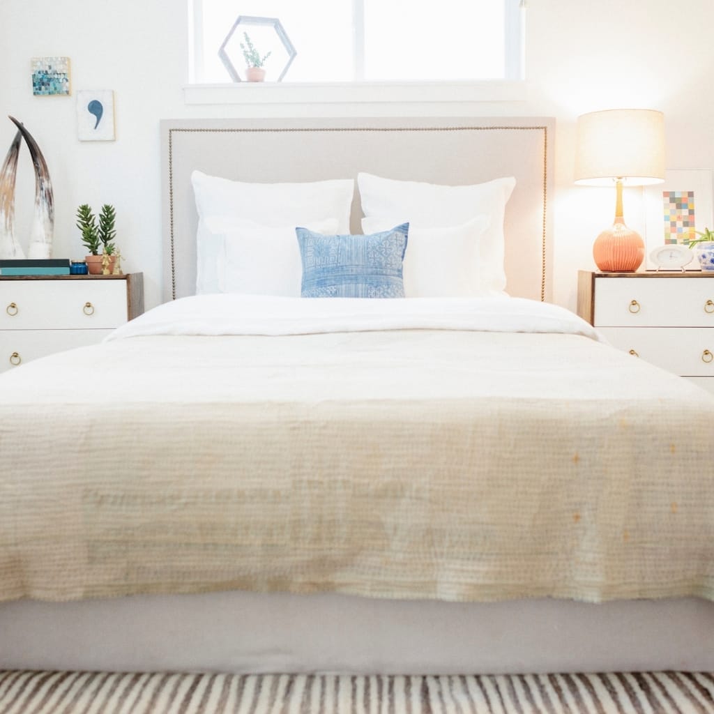 A neatly made bed with white pillows and a small blue decorative pillow in the center. The headboard is light-colored, and there are white nightstands on both sides. The left nightstand has small potted plants and decorative items, while the right side features a lamp with a pink base and some decor items. Behind the bed, there is a window with sunlight streaming in, illuminating the room.